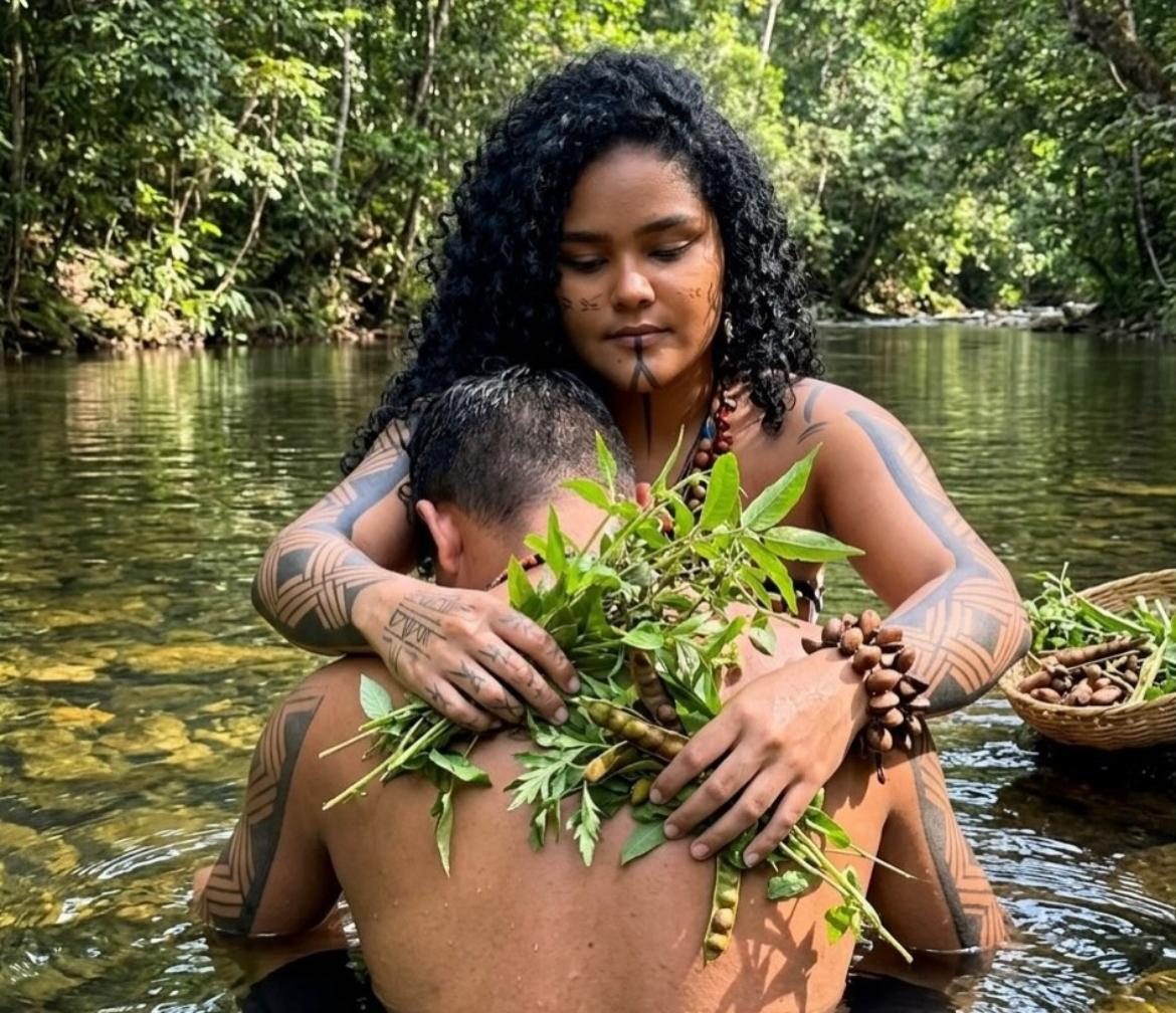A-yá dentro de um rio na floresta, abraçando por trás uma pessoa e cobrindo seus ombros com ramos de ervas durante um banho ritual